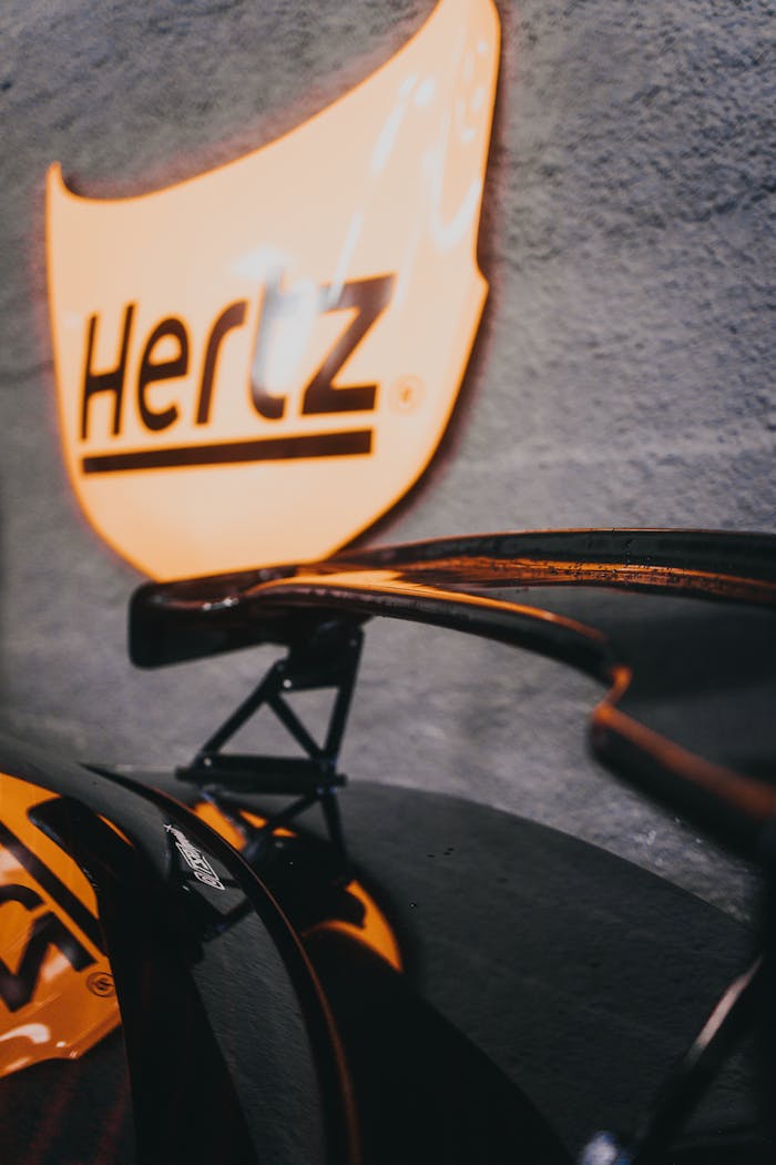 Close-up of a car spoiler with a glowing Hertz sign in the background.